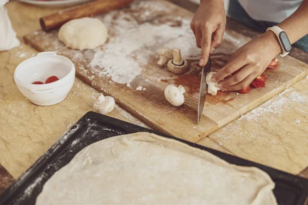 Kadınların elleri rulo hamurla birlikte bir yemek tepsisinin yanında tahta bir tahtanın üzerinde pizza mantarlarını keser. 