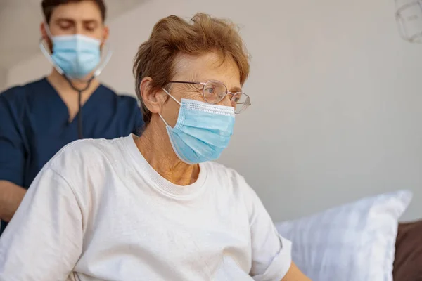 Doctor in face mask listening to old womans patient breathing, using stethoscope in hospital ward