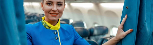 Cheerful flight attendant in air hostess uniform looking at camera and smiling while holding curtains in airplane