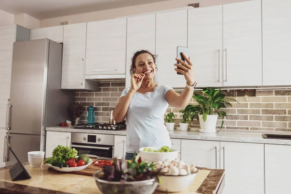 A woman standing in the kitchen in front of the table with vegetables and herbs makes a selfie