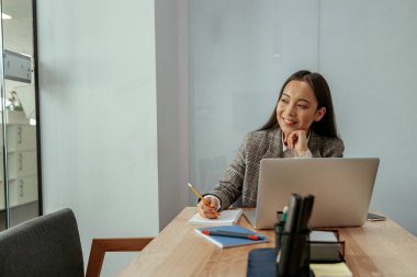 Ofiste çalışan gülümseyen Asyalı kadın bilgisayar kullanıyor ve not alıyor. Bulanık arkaplan