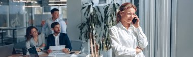 mature businesswoman is standing near window and talking on phone during meeting in modern office.