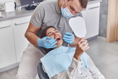 Kendine güvenen olgun doktor modern klinikte genç, mutlu, beyaz bir kadının dişlerini kontrol ediyor.