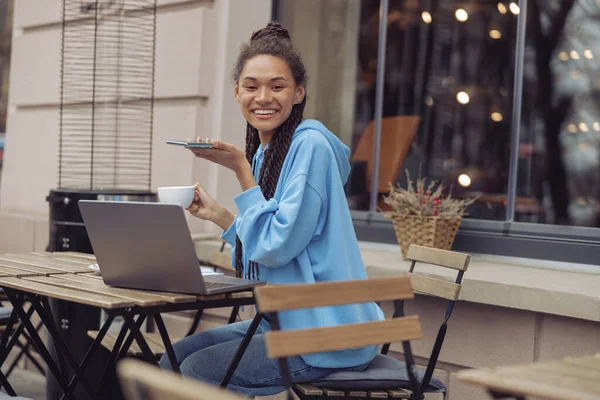 Kafe terasındaki dizüstü bilgisayarda elinde kahve ve akıllı telefonla gülümseyen güzel bir kadının portresi.