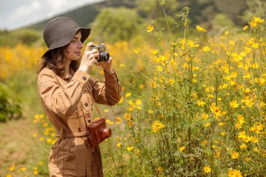 Side view of lady in hat using photo camera and taking photo of yellow flowers in Africa national park