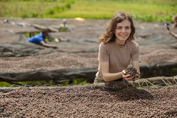 Smiling lady holding dried coffee beans in hands at farm in Africa region with workers on the background