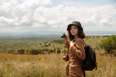 Side view of pretty lady holding photo camera and making photo while traveling through the savannah, copy space