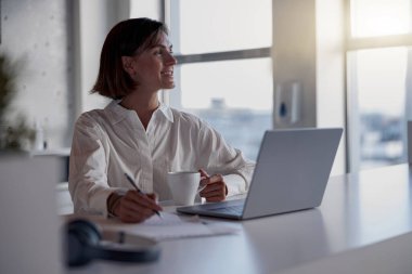 Charming businesswoman working at the office using a laptop looking away. Blurred background