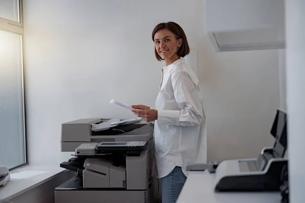 Smiling woman worker scanning a document on photocopy machine In modern office. Blurred background