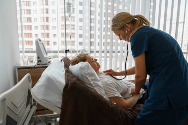 Female doctor listening to old womans patient breathing, using stethoscope in hospital ward