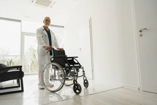 Professional senior male doctor holding wheelchair in hospital clinic hall. High quality photo