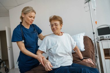 Female nurse helping patient to get up from bed in the ward at hospital. High quality photo