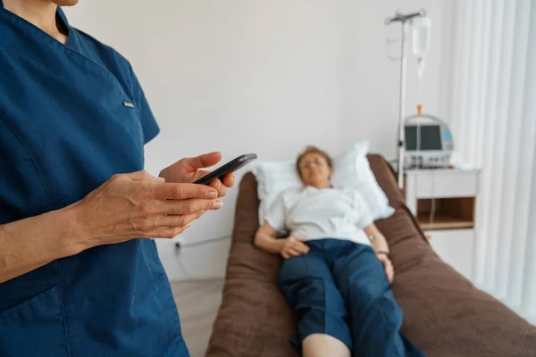 Nurse in uniform using phone standing near sick patient in hospital ward. High quality photo