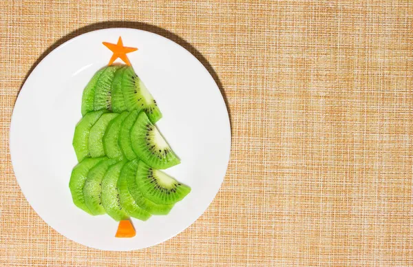 Árbol comestible de Navidad hecho de kiwi y zanahoria en plato sobre la mesa. Idea de postre para niños. Año Nuevo concepto de arte gastronómico. Vista superior, espacio de copia — Foto de Stock
