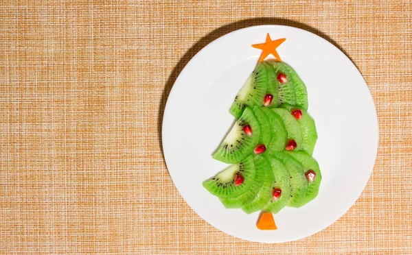 Árvore comestível de Natal feita de kiwi, cenoura e romã na placa na mesa. Ideia de sobremesa para crianças. Ano Novo conceito de arte alimentar. Vista superior, espaço de cópia — Fotografia de Stock