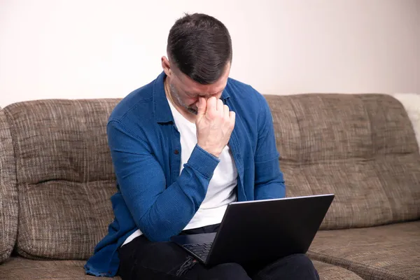 El hombre molesto con exceso de trabajo que masajea el puente de la nariz, sufre dolor de cabeza, fatiga visual después de mucho tiempo trabajando con el ordenador portátil, sentado en el sofá — Foto de Stock