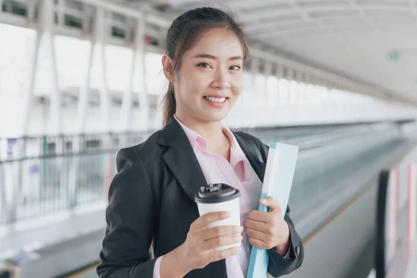 young Asian businesswoman in black suit holding folder and cup of coffee and standing on walkway station.