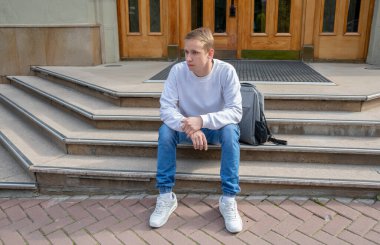 20-25 yaşlarında, yaz kıyafetleri içinde sırt çantalı genç bir adam enstitünün girişindeki basamaklarda oturuyor. Kapalı giriş kapıları. Konsept: İşten kovuldum, derslere geç kaldım, cevap bekliyorum.