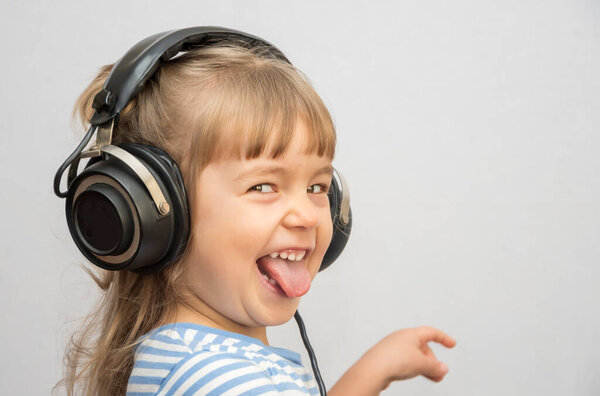 A cheerful girl in headphones shows her tongue and listens to music on a light background. Concept: learning a foreign language, old headphones sound equipment.