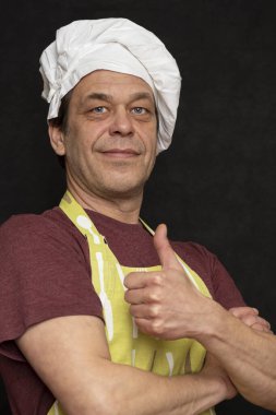 45-50 yaşlarında, beyaz şapkalı, siyah önlüklü yaşlı bir aşçının portresi. Parmağını kaldırıp kameraya bakıyor.