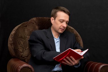 Bir adam koltuğa oturur ve bir deftere bir şeyler yazar, dikkatle dinler ve her şeye burnunu sokan, karanlık arka plana bakar..