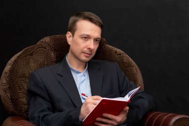 Bir adam koltuğa oturur ve bir deftere bir şeyler yazar, dikkatle dinler ve her şeye burnunu sokan, karanlık arka plana bakar..