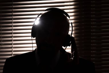 Bir FBI gizli ajanı kulaklıkla dinler ve pencerenin arka planına karşı bir konuşma kaydeder. Panjurlar, silüet aydınlatma, seçici odak, koyu tonlama..
