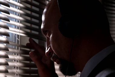 Gizli bir ajan, kulaklıklarla yapılan bir konuşmayı dinliyor. Pencerenin perdelerinden sokağa bakıyor. Silüet aydınlatma, seçici odaklanma, karanlık ton..