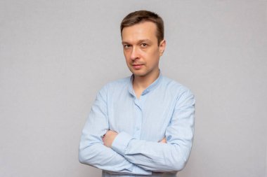 Açık renk tişörtlü, gülümseyen esmer, kameraya bakıyor, kollarını kavuşturuyor, gri bir arka planda..