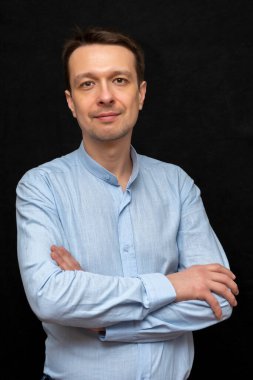 30-35 yaşlarında, kollarını kavuşturmuş mavi gömlekli bir adamın portresi siyah bir arka planda duruyor. Belki de avukat, koç ya da psikologtur..