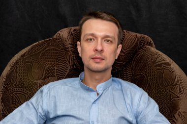 Mavi gömlekli, siyah arka planda koltukta oturan 30-35 yaşlarında bir adamın portresi. Belki de avukat, koç ya da psikologtur..