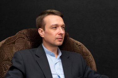 Resmi giyinmiş genç bir adam siyah arka planda bir koltukta oturuyor ve başka tarafa bakıyor..