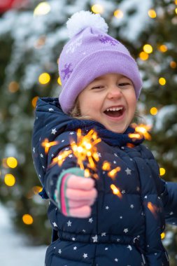 Mutlu küçük bir kız elinde yanan bir maytap tutuyor. Bir Noel ağacının arka planında. Yakın plan, seçici odaklanma. Kavram: şenlik havası, Noel ve yeni yıl, çocukların neşesi.