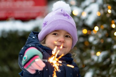 Mutlu küçük bir kız elinde yanan bir maytap tutuyor. Bir Noel ağacının arka planında. Yakın plan, seçici odaklanma. Kavram: şenlik havası, Noel ve yeni yıl, çocukların neşesi.