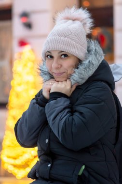 Kırk yaşındaki şapkalı ve kışları sokakta kalın ceketli bir kadının portresi şehir yılbaşı dekorasyonlarının arka planına karşıdır..