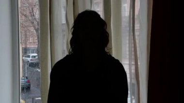 Arka plandaki pencerenin arka planında genç bir kadın bir hikaye anlatır, yüzü göremezsiniz, seçici odaklanma, karanlık ton, yakın çekim.