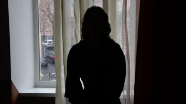 Genç bir kadının silueti arka plandaki bir pencerenin arka planında, yüzü görünür değil, seçici odaklanma, karanlık ton, orta plan.