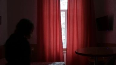 Genç bir kadının silueti, perdeleri karanlık bir odadaki pencerenin arka planına doğru açar. Yüzü görünmez, seçici odak noktası, koyu renk tonu yoktur..