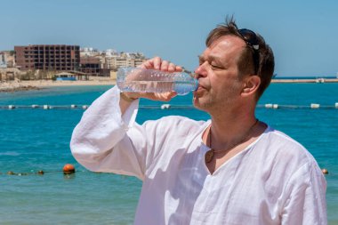 45-50 yaşlarında yaşlı bir adam denizin arka planına karşı plastik bir şişeden su içiyor. Kavram: Vücudun susuzluğu ve susuzluğu, hayat veren nem.