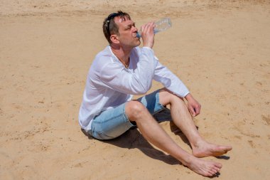 45-50 yaşlarında yaşlı bir adam çölde kumların üzerinde duran plastik bir şişeden açgözlülükle su içiyor. Kavram: susuzluk ve susuzluk, hayat veren nem, sıcak ülkelerde turizm.