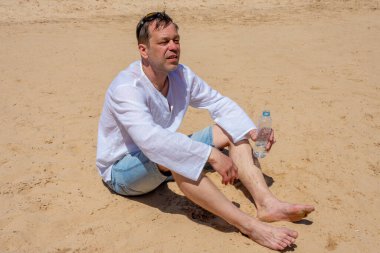 Sıcaktan bitap düşmüş, 45-50 yaşlarında yaşlı bir adam kumda oturuyor ve elinde plastik bir şişe su tutuyor. Kavram: susuzluk ve susuzluk, hayat veren nem, sıcak ülkelerde turizm.