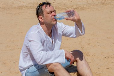 45-50 yaşlarında yaşlı bir adam çölde kumların üzerinde duran plastik bir şişeden açgözlülükle su içiyor. Kavram: susuzluk ve susuzluk, hayat veren nem, sıcak ülkelerde turizm.