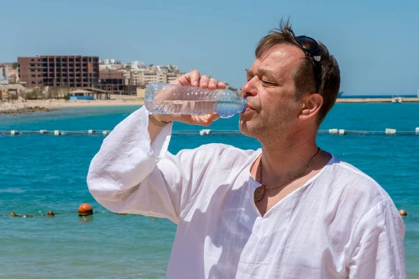 45-50 yaşlarında yaşlı bir adam denizin arka planına karşı plastik bir şişeden su içiyor. Kavram: Vücudun susuzluğu ve susuzluğu, hayat veren nem.