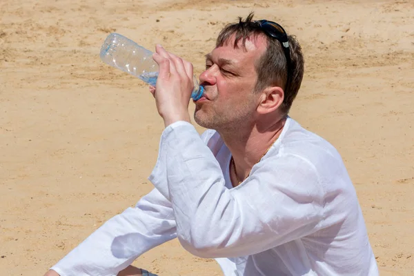 45-50 yaşlarında yaşlı bir adam çölde kumların üzerinde duran plastik bir şişeden açgözlülükle su içiyor. Kavram: susuzluk ve susuzluk, hayat veren nem, sıcak ülkelerde turizm.