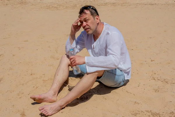 Sıcaktan bitap düşmüş, 45-50 yaşlarında yaşlı bir adam kumda oturuyor ve elinde plastik bir şişe su tutuyor. Kavram: susuzluk ve susuzluk, hayat veren nem, sıcak ülkelerde turizm.