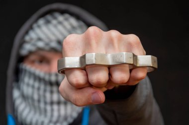 Yüzü koyu bir arka planda, seçici bir odak noktasında eşarplı bir adamın elinde pirinç muşta. Kavram: holiganların sokak kavgaları, vatandaşlara saldırılar, kentsel suçlar.