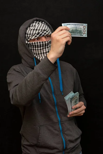 一个穿着连帽衫的男人看着一张100美元的钞票 他的脸上布满了阿拉伯围巾 深色的背景和选择性的焦点 — 图库照片