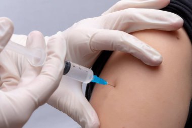Doktor aşıyı arka planda, yakın planda bir kızın omzuna enjekte ediyor..