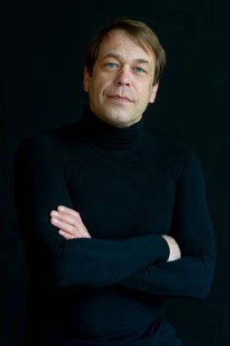 45-50 yaşlarında, siyah arka planda golf kazağı giyen, kameraya bakan ciddi bir adamın portresi. Belki de eski bir asker, iş adamı, avukat ya da karizmatik bir aktördür..