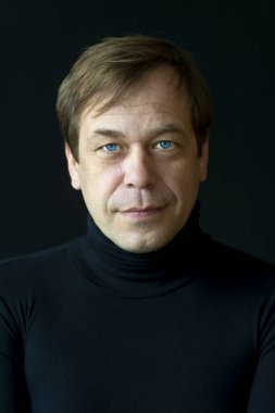 45-50 yaşlarında, siyah arka planda golf kazağı giyen, kameraya bakan ciddi bir adamın portresi. Belki de eski bir asker, iş adamı, avukat ya da karizmatik bir aktördür..